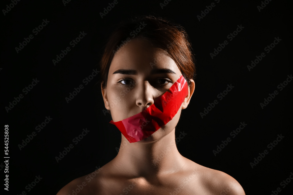 Young woman with taped mouth on dark background. Censorship concept