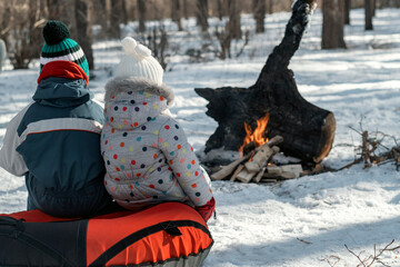 two young children in warm jackets sit in the winter forest and look at fire. back view. weekends or