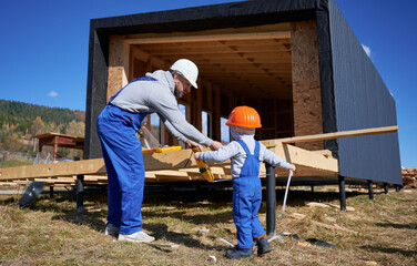 Wall Mural - Father with toddler son building wooden frame house. Male builder and kid playing with tape measure on construction site, wearing helmet and blue overalls on sunny day. Carpentry and family concept.