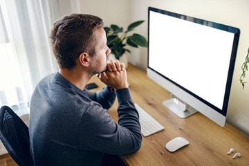a focused young programmer is sitting at the home office and looking at the code on monitor.