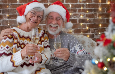 Cheerful senior adult couple at home enjoying christmas together in december wearing santa hats. Man and woman toasting with sparkling wine glasses looking at camera in holiday leisure. Xmas tree