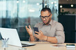 © Liubomir - African american man received bad news notification online from phone, businessman in shirt working with laptop in middle of modern office building, man in glasses reading online letter.