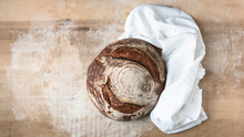 Single Round Loaf Of Crusty Baked Sourdough Bread On Lightly Floured Blonde Wood Board With White Dish Towel. 
