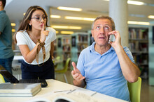 Disappointed Latin Woman Making Silence Gesture To Mature Male, While He Is Using Laptop And Talking On Mobile Phone In The Library. High Quality Photo