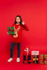 Wall Mural - full length of happy child in knitted sweater showing thumb up near Christmas presents on red