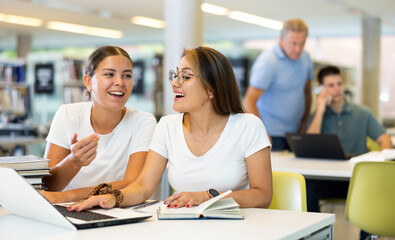 Canvas Print - Two friendly young adult female students studying in university library, using books and laptop while preparing for lecture