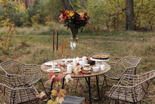 Cozy Autumn Picnic In The Park. Festive Setting Table Decorated Wildflowers In Vase, Candles, Food, Hot Drinks, Maples Leaves And Wicker Chairs Outdoors. Straw Baskets And Pumpkins On The Grass