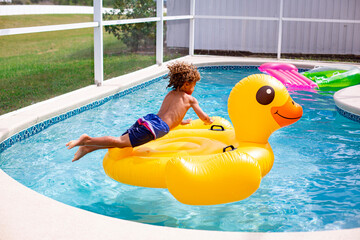 Young diverse little boy jumping onto a large inflatable pool toy in a backyard swimming pool on a warm summer day. Action photo of the child jumping through the air