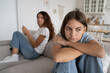 © DimaBerlin - Thoughtful Polish school age girl leans on knees looks into distance feels sad because of house arrest or quarantine. Casual worried Spanish woman sits on sofa behind daughter for teen issues concept