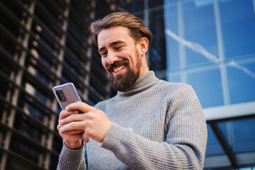 Happy business young male enjoying using a mobile phone app sharing media in internet. Awesome man texting and laughing with cellphone outdoors. High quality photo