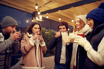 Happy smiling friends with cups of mulled wine having fun, spending time together at winter fair at evening time. Winter holidays, Christmas concept