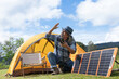 Senior camper man checking solar panels to match the direction of sunlight in front of his yellow tent, man holding smartphone and charging from solar panels mobile in summer camp