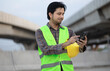 © anut21ng Stock - man texting message on cellphone, Asian construction worker wearing a helmet stands at construction site touching smartphone