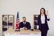© Studio Romantic - Young male judge sit at desk in court, female witness oath on constitution before trial. Man juror or prosecutor against American flag in law. Justice and jurisdiction concept.