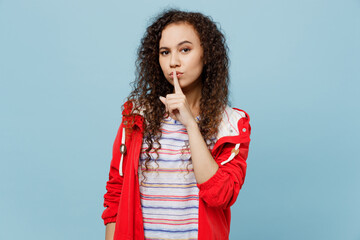 Wall Mural - Young secret woman of African American ethnicity wear red jacket say hush be quiet with finger on lips shhh gesture isolated on plain pastel light blue cyan background Wet fall weather season concept