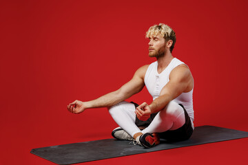 Full body young strong sporty sportsman man wear white clothes spend time in home gym sit on yoga mat meditate hold hands in om gesture isolated on plain red background Workout sport fit body concept