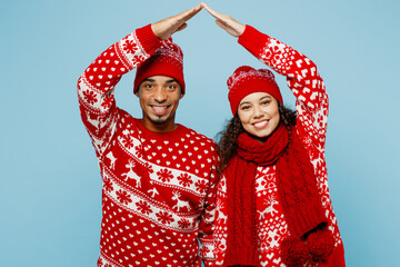 Merry young couple two man woman wear red Christmas sweater Santa hat posing hold folded hands above head like house roof isolated on plain pastel blue background. Happy New Year 2023 holiday concept.