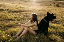 Young Brunette Woman Sitting In A Field Next To A Big Black Dog Giant Schnauzer Breed.