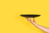 Fototapeta Kawa jest smaczna - Mano de mujer sosteneniendo un plato negro sobre un fondo amarillo brillante liso y aislado. Vista de frente y de cerca. Copy space