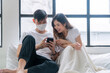© kaew6566 - Joyful asian young man wear mask with wife use mobile phone watching movie or shopping online together relax on bed. Asian young couple browsing internet smartphone. Family lifestyle technology online
