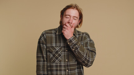 Tired handsome bearded man in checkered shirt yawning sleepy inattentive feeling somnolent lazy bored gaping suffering from lack of sleep. Young blonde guy boy isolated on beige studio background