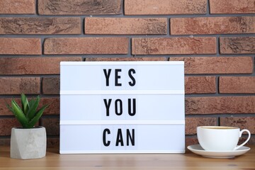 Wall Mural - Lightbox with phrase Yes You Can, cup of coffee and potted houseplant on table against brick wall. Motivational quote