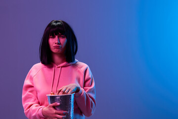 Wall Mural - Confused unhappy awesome brunet woman in pink hoodie eat popcorn squinting look at camera posing isolated in blue violet color light studio background. Neon party Cyberpunk Cinema concept. Copy space
