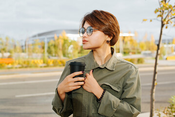 Wall Mural - Portrait of abeautiful stylish young asian woman with short haircut and glasses holding coffee standing outdoors on sunny evening and looking away