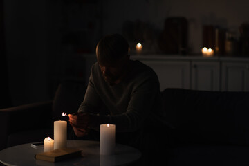 Wall Mural - man lighting candles in dark kitchen near mobile phone and book during energy blackout.