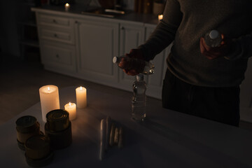 Wall Mural - cropped view of man with bottled water near table with canned food and candles in dark kitchen.