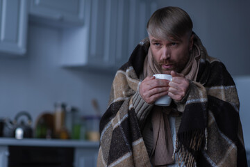 Wall Mural - man standing under warm blanket and blowing at hot tea during power outage at home.