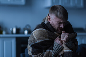 Wall Mural - frozen man with closed eyes sitting in warm blanket during energy blackout at home.