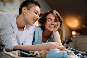 Wall Mural - Two lesbian girls smiling and holding photo while lying in bed at home