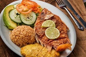 Top view of a white plate with Pescado Frito, rice, and a vegetable salad on a wooden table