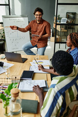 Wall Mural - Confident coach explaining financial graph to young African American colleagues sitting by desk in front of him at business seminar