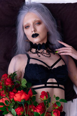 Dark goth girl in underwear laying on her bed, holding roses and seducing you