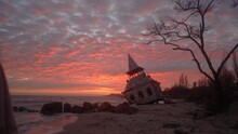 Woman Stands, Looks At Mysterious Old Flooded Leaning Wooden House, Chapel Washed By Sea Waves, Sinking In Water On Beach. Girl Goes Away After Visiting Damaged Abandoned Home On Vivid Pink Sunset.
