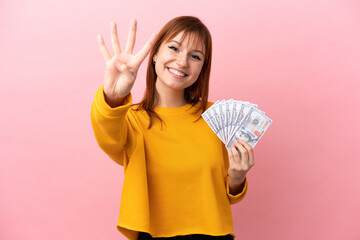 Redhead girl taking a lot of money isolated on pink background happy and counting four with fingers