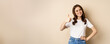 © Mix and Match Studio - Portrait of modern young woman showing thumbs up, like and approve, smiling pleased, recommending company or website, standing over beige background