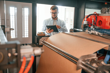 Holding digital tablet when standing near sheet cutting machine. Print house worker in white clothes is indoors