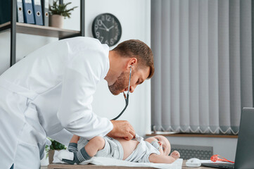 Wall Mural - Using stethoscope. Doctor in white coat is taking care of little toddler indoors