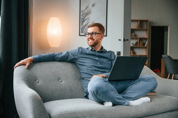 Wall Mural - Sitting and looking at the side. Male freelance worker is using laptop at home. Cozy apartment