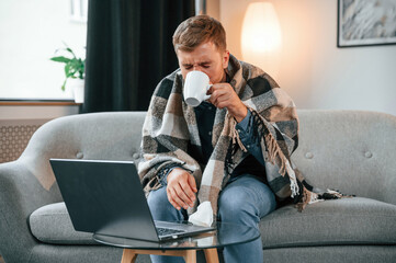 Wall Mural - Drinking a tea. Sick man is sitting on the sofa at home by the laptop. Conception of illness