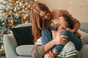 Wall Mural - Woman is on the sofa, embracing her man by the legs. Lovely young couple are celebrating New Year at home