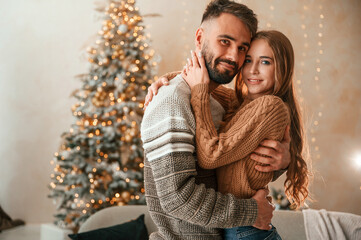 Wall Mural - Standing against the fir with garlands on it. Lovely young couple are celebrating New Year at home