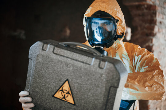 Blue light is going from hard case that is with hazard sign. Man dressed in chemical protection suit in the ruins of the post apocalyptic building
