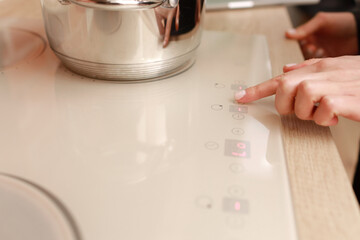 Female hand turns on electric hob closeup, finger presses sensor button on modern white induction cooker. Metal steel saucepan on kitchen stove top panel, modes of cooking surface, Domestic equipment