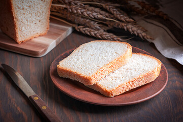 Wall Mural - Homemade sliced loaf of bread made of wheat flour and yeast served on plate on dark brown wooden table with ripe grain crop, knife and towel at kitchen for morning breakfast as healthy baked snack