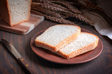 Poster - Homemade sliced loaf of bread made of wheat flour and yeast served on plate on dark brown wooden table with ripe grain crop, knife and towel at kitchen for morning breakfast as healthy baked snack