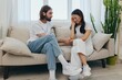 © SHOTPRIME STUDIO - A sad Asian woman talks to a man in tears at home sitting on the couch. Young couple of different nationalities and conflict of interest in a couple